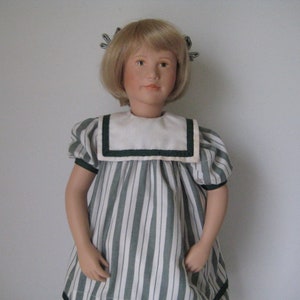 Peut inclure: Une poup&eacute;e en porcelaine aux cheveux blonds portant une robe &agrave; rayures vertes et blanches avec un col blanc et une bordure verte.