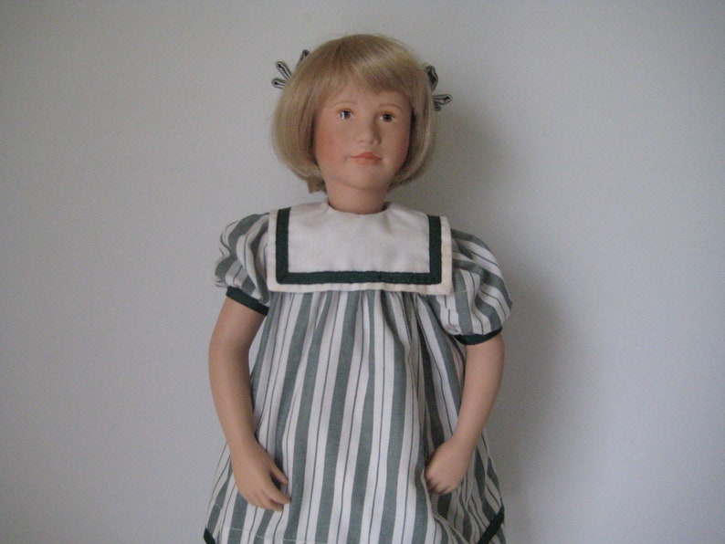 Peut inclure: Une poup&eacute;e en porcelaine aux cheveux blonds portant une robe &agrave; rayures vertes et blanches avec un col blanc et une bordure verte.