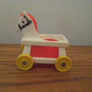 May include: A vintage toy horse-drawn cart featuring a white horse head with a red mane and black bridle details. The cart body is white with a red centre panel and is supported by four yellow plastic wheels with small silver-coloured metal hubs.
