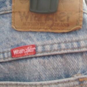 Op de afbeelding: Close-up van een lichtblauwe spijkerbroek met een bruine leren patch en een rood Wrangler-label. De leren patch is voorzien van het Wrangler-logo en de tekst "AUTHENTIC JEANS". Het rode label is op de spijkerbroek gestikt.