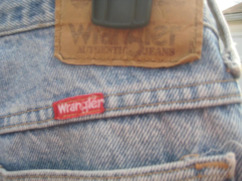 Op de afbeelding: Close-up van een lichtblauwe spijkerbroek met een bruine leren patch en een rood Wrangler-label. De leren patch is voorzien van het Wrangler-logo en de tekst "AUTHENTIC JEANS". Het rode label is op de spijkerbroek gestikt.