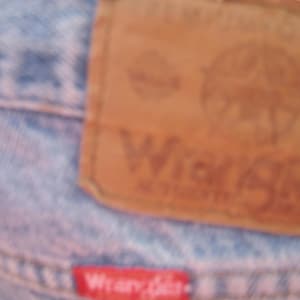 Op de afbeelding: Close-up van een lichtblauwe spijkerbroek met een bruin leren label. Het label bevat het woord "Wrangler" en een logo. Een klein rood label met het woord "Wrangler" is ook zichtbaar.