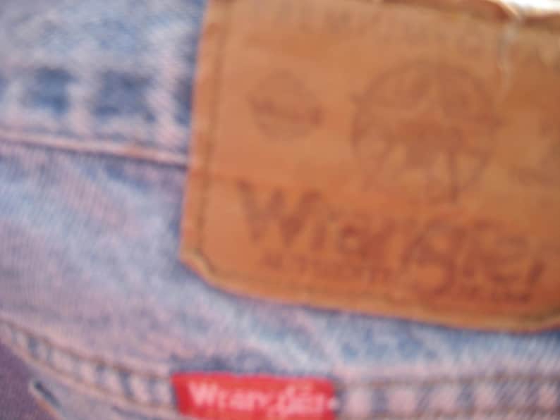 Op de afbeelding: Close-up van een lichtblauwe spijkerbroek met een bruin leren label. Het label bevat het woord "Wrangler" en een logo. Een klein rood label met het woord "Wrangler" is ook zichtbaar.