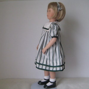 Peut inclure: Une poup&eacute;e en porcelaine portant une robe &agrave; rayures blanches et vertes avec un col marin blanc et un ruban vert dans les cheveux. Elle porte des chaussures noires et des chaussettes blanches.
