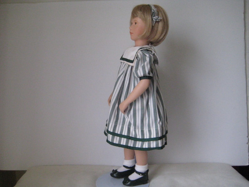 Peut inclure: Une poup&eacute;e en porcelaine portant une robe &agrave; rayures blanches et vertes avec un col marin blanc et un ruban vert dans les cheveux. Elle porte des chaussures noires et des chaussettes blanches.