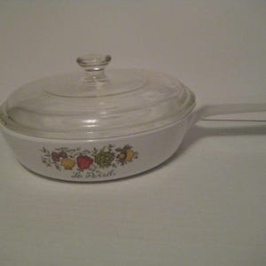 May include: White casserole dish with a clear glass lid and a long handle. The dish features a colorful fruit and vegetable design around the rim, with the text "Le Persil" below the design. The lid has a small knob.