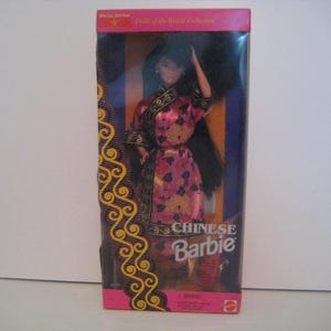Puede incluir: Una muñeca Barbie china con un vestido tradicional rosa y dorado, exhibida en su caja original. La caja es rosa con detalles dorados y se ven las palabras "Chinese Barbie".