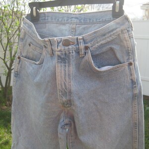 Op de afbeelding: Lichtblauwe spijkerbroek van denim die aan een zwarte hanger hangt. De jeans hebben een klassiek ontwerp met vijf zakken met een knoop- en ritssluiting. De denim heeft een vervaagde wassing, met lichtere plekken rond de zakken en naden.