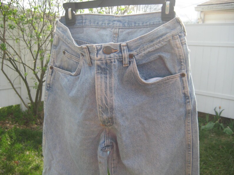 Op de afbeelding: Lichtblauwe spijkerbroek van denim die aan een zwarte hanger hangt. De jeans hebben een klassiek ontwerp met vijf zakken met een knoop- en ritssluiting. De denim heeft een vervaagde wassing, met lichtere plekken rond de zakken en naden.