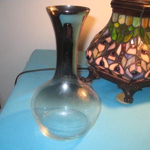 May include: A clear glass vase with a bulbous base and a tall, slender neck that flares slightly at the top. The glass features a subtle gradient, transitioning from clear at the bottom to a darker, reflective metallic finish near the rim.