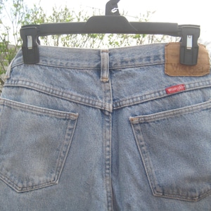 Op de afbeelding: Lichtblauwe denim jeans met twee achterzakken en een bruine leren patch. Het rode Wrangler-label is zichtbaar op de achterzak. De jeans hangt aan een zwarte hanger.