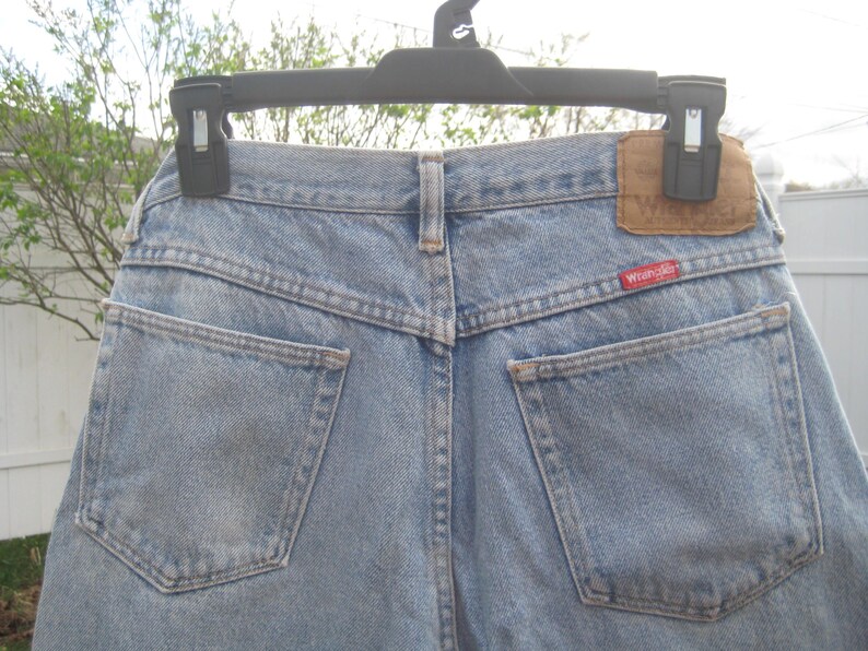 Op de afbeelding: Lichtblauwe denim jeans met twee achterzakken en een bruine leren patch. Het rode Wrangler-label is zichtbaar op de achterzak. De jeans hangt aan een zwarte hanger.