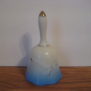 Puede incluir: Una campana de porcelana blanca con un diseño de gaviota azul y blanca. La campana tiene un asa dorada.