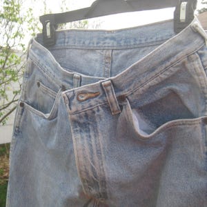 Op de afbeelding: Lichtblauwe spijkerbroek hangend aan een zwarte hanger. De jeans heeft voor- en achterzakken, een knoopsluiting en riemlussen. De denim heeft een vervaagde uitstraling.