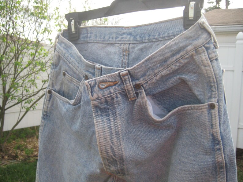 Op de afbeelding: Lichtblauwe spijkerbroek hangend aan een zwarte hanger. De jeans heeft voor- en achterzakken, een knoopsluiting en riemlussen. De denim heeft een vervaagde uitstraling.