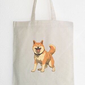 Puede incluir: Una bolsa de tela blanca con una imagen de dibujos animados de un perro Shiba Inu con un pelaje marrón y blanco. El perro está sonriendo con la boca abierta y la lengua fuera. El perro lleva un collar negro con una etiqueta plateada.