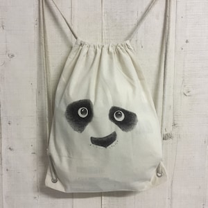 Könnte beinhalten: Eine weiße Kordeltasche mit einem schwarzen Pandagesicht, das auf der Vorderseite aufgedruckt ist. Der Panda hat schwarze Augen und eine schwarze Nase und einen schwarzen Mund.