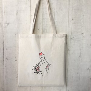 May include: A white canvas tote bag with a pink heart and hand gesture design. The hand gesture is a Korean heart sign, also known as the "mini heart".
