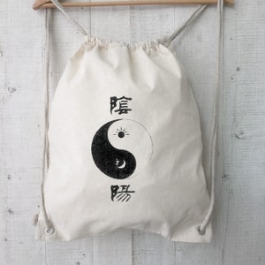 Può includere: Una borsa a cordoncino bianca con un simbolo yin yang nero e caratteri cinesi. La borsa è realizzata in tela e ha un design semplice.