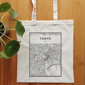 May include: A white canvas tote bag with black handles and a black and white map of Tokyo, Japan printed on the front. The map is surrounded by a black border and the text "Tokyo, Japan" is printed above the map.