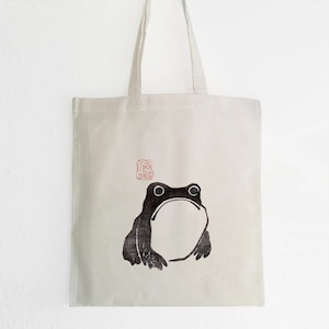 Peut inclure: Un sac fourre-tout blanc avec une illustration noire et blanche d'une grenouille. La grenouille est assise sur le sac et a un tampon rouge au-dessus qui dit "吉" en japonais.