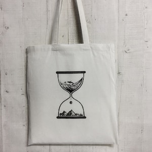 May include: A white tote bag with black handles and a black hourglass design. The hourglass is filled with waves and a small whale. The design is centered on the bag.