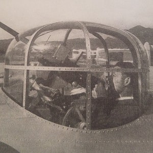 May include: A black and white photo of a machine gun turret mounted on top of an airplane fuselage. The turret is made of metal and has a clear dome. A person is sitting inside the turret, aiming the gun.
