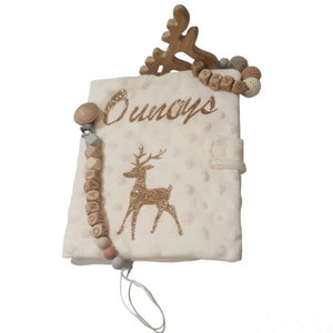 May include: Cream baby book with a gold glitter deer design and the name "Ounays" in gold glitter. A wooden deer head and a pacifier clip with wooden beads are attached. The pacifier clip has the name "Ounays" spelled out on it.