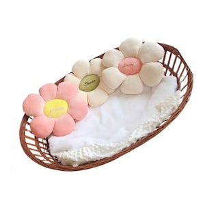 Peut inclure: Trois coussins en peluche en forme de fleur dans un panier en osier. Les coussins sont en rose, blanc et vert avec des noms brodés "Olivia", "Savannah" et "Love".