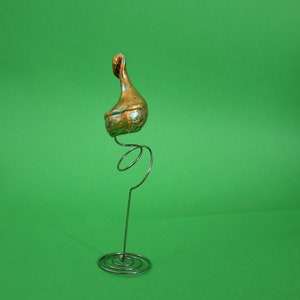 Könnte beinhalten: Eine goldfarbene Keramik-Vogelfigur, die auf einem Metallfederständer sitzt. Der Ständer hat eine runde Basis.