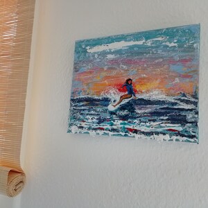 Könnte beinhalten: Ein Gemälde, das eine Person beim Surfen auf einer Welle zeigt. Das Gemälde ist in einem farbenfrohen Stil mit Blautönen, Rosa und Orange gehalten. Die Person trägt einen blauen Neoprenanzug und reitet auf einem weißen Surfbrett.