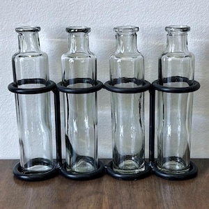 May include: A set of four clear glass bottles held in a black metal frame. Each bottle features a slender neck and a cylindrical body. The metal frame consists of four circular rings connected by vertical bars, designed to hold the bottles upright.