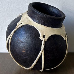 May include: A round, dark charcoal-colored vessel with a wide opening and a textured surface. The pot is encased in a decorative network of light cream-colored leather strips that wrap around the body and secure near the neck.