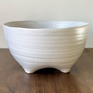 May include: A white ceramic bowl with a textured, ridged exterior and three small feet. The bowl has a wide, slightly irregular rim and a smooth, glossy finish. The bowl is sitting on a wooden surface.