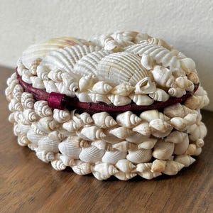 May include: A white ceramic mug with a black handle and the text "But First, Coffee" in a bold, black font. The mug is set against a blurred background, suggesting a cozy setting. The mug appears to be approximately 4 inches tall.