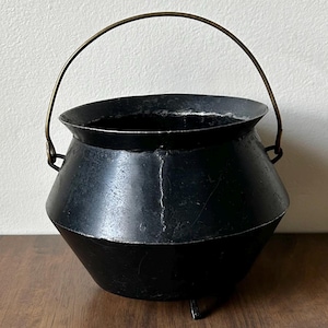 May include: A black cauldron with a brass handle and three small feet. The cauldron has a rounded body and a wide opening. The handle arches over the top, and the cauldron sits on a wooden surface.