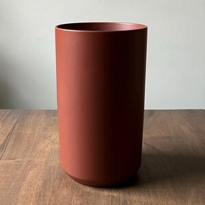 May include: A terracotta colored cylindrical ceramic vase with a smooth, matte finish.