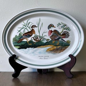 May include: An oval ceramic serving platter featuring a colorful illustration of wood ducks in a natural habitat. The text WOOD DUCK Anas Sponsa is printed below the birds. The platter has a green rim and rests on a dark wooden stand.