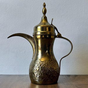 May include: A brass coffee pot featuring a long, curved spout, a tall lid with a pointed finial, and a slender handle. The lower body of the pot is decorated with an intricate, embossed pattern of geometric shapes and floral motifs.