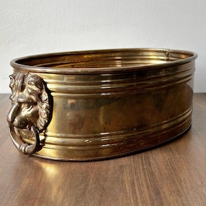 May include: Oval brass planter with a lion's head handle. The planter has a ridged design and a polished finish. The lion's head handle is detailed and adds a decorative element. The planter is suitable for indoor or outdoor use.