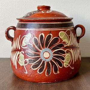May include: A red ceramic jar with a lid, featuring a floral design in black, white, and green. The jar has two handles and is set on a wooden surface. The lid has a small knob and a dark brown band.