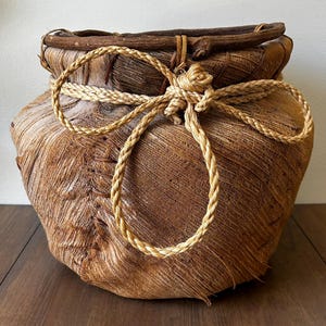 May include: A round, textured brown basket crafted from fibrous plant material. The rim is reinforced with a dark, thick wooden branch. A light tan, braided rope is tied in a decorative bow around the upper section of the basket body.