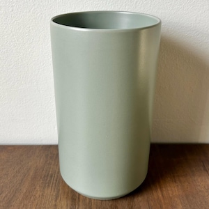 May include: A tall, cylindrical, sage green ceramic vase. The vase has a smooth, matte finish and a rounded base. The interior is the same color as the exterior. The vase is displayed on a dark wood surface.