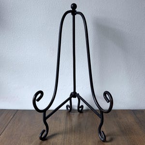 May include: A black metal display stand featuring a tall, arched frame with a small spherical finial at the top. The base consists of three curved legs with scrolled ends, designed to hold items upright with a central support rod and two front arms.