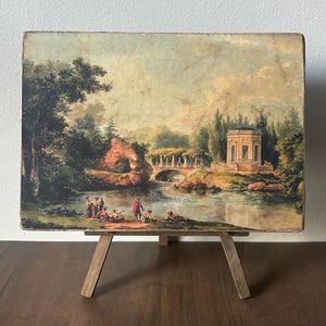 May include: An antique landscape painting on a wooden panel, depicting a classical scene with a bridge, a temple, and figures by a lake. The artwork is displayed on a small wooden easel, showcasing the aged appearance of the painting.