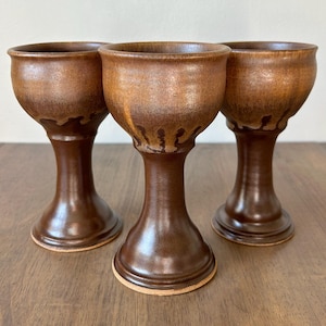 May include: Three handmade brown ceramic goblets with a glossy finish. The goblets have a wide rim, a slender stem, and a round base. The glaze has a dripped effect near the rim. The goblets are displayed on a wooden surface.