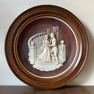 May include: A round, framed wall plaque featuring a bas-relief scene of a couple on a staircase. The relief is off-white, set against a dark brown background. The frame is made of wood with a gold inner rim. The plaque is signed "C. Romanelli".