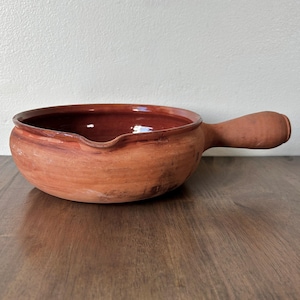 May include: A rustic, terracotta-colored ceramic bowl with a handle. The bowl has a wide, shallow design with a dark red interior and a small spout. The handle is rounded and extends from the side of the bowl. The bowl is likely handmade.