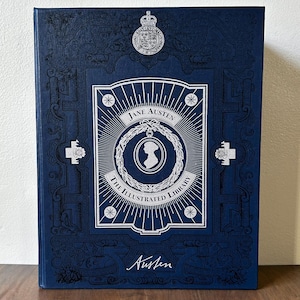 May include: A hardcover book titled "Jane Austen The Illustrated Library" in navy blue. The cover features a white illustration of a woman's silhouette within a circular design, surrounded by radiating lines. The book has decorative floral and geometric patterns.