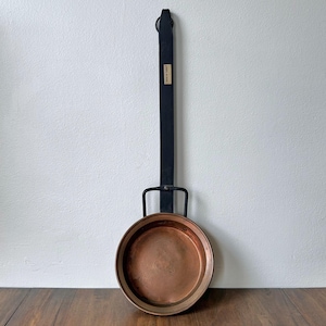 May include: A copper pan with a long, black handle and a leather loop for hanging. The pan has a circular cooking surface and a dark rim. A small, rectangular tag is attached to the handle.